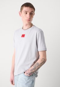 T-shirt in cotone grigio chiaro con un patch logo rosso sul petto, abbinato a jeans azzurri, con un tatuaggio visibile sul braccio.