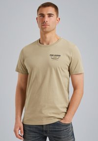 Beige bavlněné tričko s krátkým rukávem, kulatým výstřihem a potiskem na levé části hrudi. Nosené s modrými džínami.