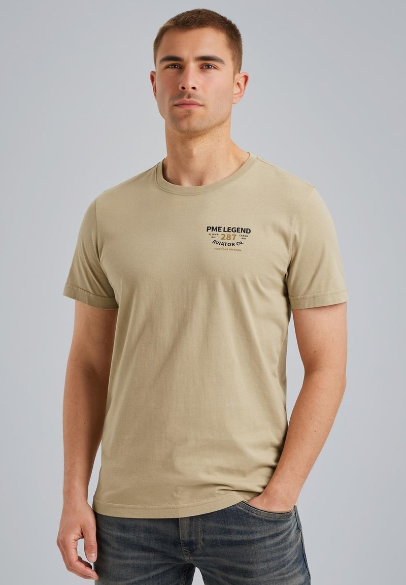 Beige bavlněné tričko s krátkým rukávem, kulatým výstřihem a potiskem na levé části hrudi. Nosené s modrými džínami.