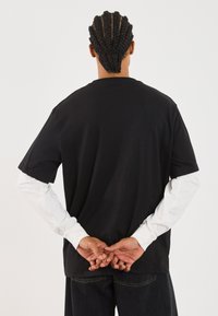 Sort oversized t-shirt med korte ærmer, båret over en hvid langærmet skjorte. Bagfra, afslappet pasform, bomuldsmateriale, ensfarvet design.