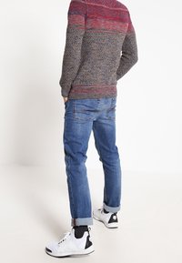 Mann trägt einen mehrfarbigen Strickpullover, hochgekrempelte blaue Jeans, schwarze Socken und weiße High-Top-Sneaker, steht mit den Händen in den Taschen.