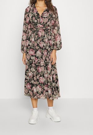 Femme portant une robe midi noire avec un imprimé floral rose et vert, des manches longues transparentes, et des baskets blanches avec des chaussettes blanches montantes.