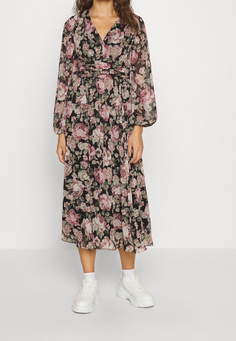 Femme portant une robe midi noire avec un imprimé floral rose et vert, des manches longues transparentes, et des baskets blanches avec des chaussettes blanches montantes.