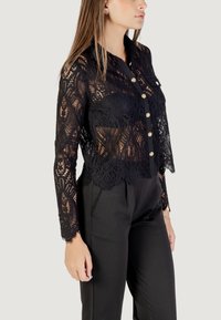 Blusa nera in pizzo con intricati motivi floreali, maniche lunghe, colletto e chiusura a bottoni, abbinata a pantaloni neri.