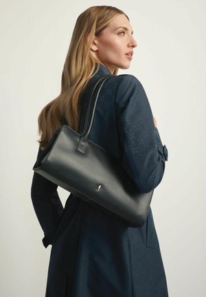 Kobieta z długimi blond włosami, ubrana w ciemnoniebieski wzorzysty płaszcz, niosąca elegancką czarną skórzaną torebkę na ramieniu.