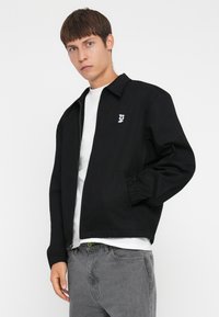 Svart zip-up jacka med krage, elastiska ärmslut och en liten logotyp på vänster bröst. Bärs över en vit t-shirt och grå jeans.