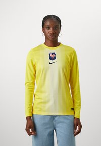 Ilgi piedurkņu dzeltena sporta krekls ar gradienta dizainu, ar zilu emblemu ar vistas attēlu un melnu Nike logo uz krūšu.