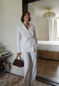La chaqueta blanca con envoltura y los pantalones a juego presentan un diseño elegante. El conjunto se complementa con un pequeño bolso marrón y un suelo texturizado.