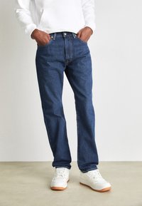 Jeans de denim azul con un corte recto, diseño de cinco bolsillos y cierre de botón. Llevados con zapatillas blancas que tienen una suela de goma.