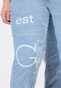 Pantalones de mezclilla azul claro con una textura arrugada que presentan texto impreso en blanco y elementos gráficos en el lado izquierdo.