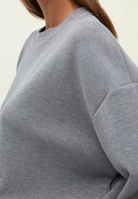 Felpa grigia realizzata in tessuto morbido, con colletto rotondo, spalle scese e una vestibilità rilassata con una superficie liscia e texture.