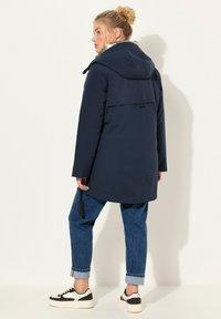 Marinblå huva med en avslappnad passform, som har en ryggslits, justerbara snören och en slät, vattenavvisande yta.