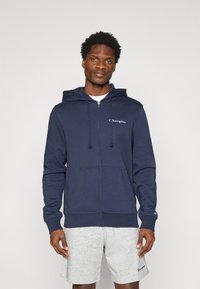 Navy Blauer Zip-Hoodie aus weichem Material, mit einer Fronttasche, Kapuze mit Kordelzug und weißem Logo auf der linken Brust. Kombiniert mit grauen Shorts.