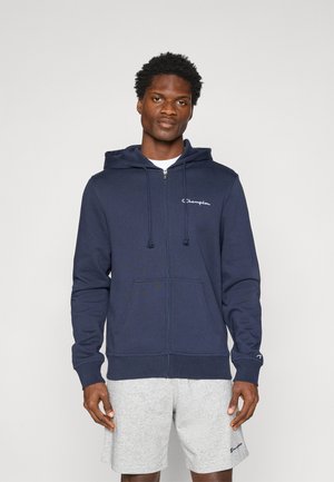 Sweat à capuche zippé bleu marine en tissu doux, avec une poche avant, une capuche à cordon et un logo blanc sur la poitrine gauche. Associé à un short gris.