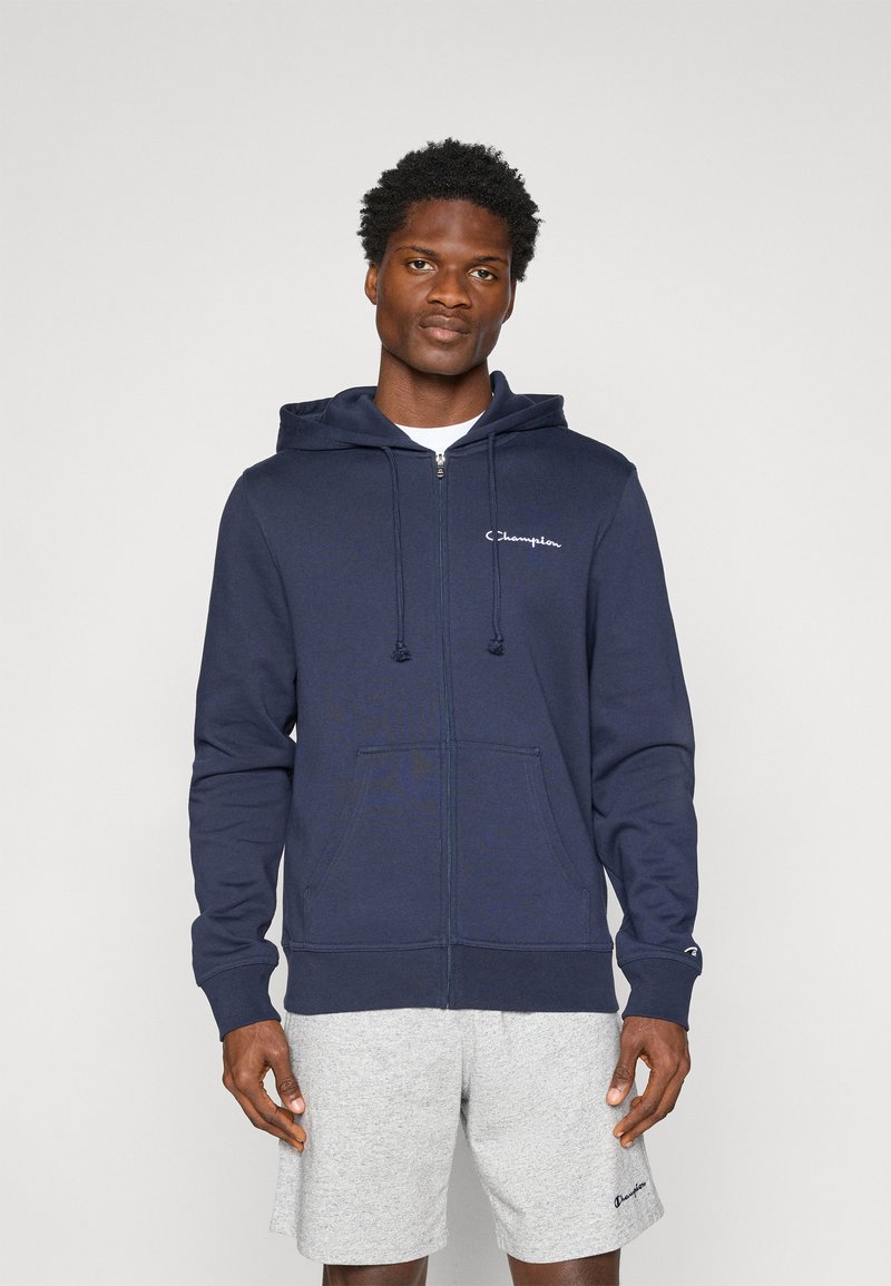 Navy Blauer Zip-Hoodie aus weichem Material, mit einer Fronttasche, Kapuze mit Kordelzug und weißem Logo auf der linken Brust. Kombiniert mit grauen Shorts.