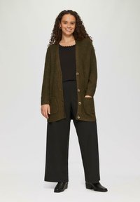 Olivgrüner Cardigan mit großen Vordertaschen, fünf Knöpfen und strukturierter Stoffqualität, kombiniert mit schwarzen, weiten Hosen und Stiefeletten.