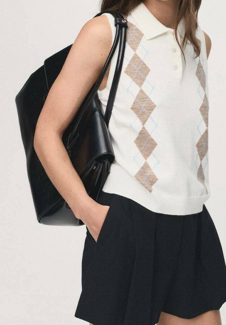 Femme portant un débardeur en maille blanche à motif losanges et un short noir, portant un grand sac à épaule en cuir noir.