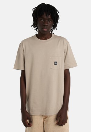 Młoda osoba o ciemnej skórze i dredach, ubrana w beżowy t-shirt z kieszenią oraz jasnobeżowe spodnie, stojąca na jednolitym tle.