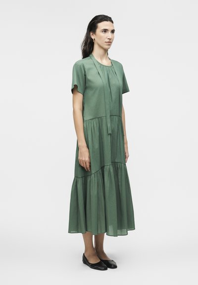 Γυναίκα που στέκεται σε ένα ρέον πράσινο μίντι φόρεμα με στρώσεις, κοντά μανίκια, λεπτομέρεια δεσίματος στον λαιμό και μαύρα επίπεδα παπούτσια πάνω σε απλό φόντο.