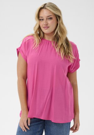 Kobieta o długich blond włosach, ubrana w jasnoróżowy krótki rękaw i niebieskie dżinsy, stojąca na tle jednolitego jasnego tła.