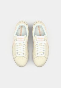 Cream-colored sneakers with white laces, perforated toe box, light blue interior lining, and subtle orange accents on the sides.