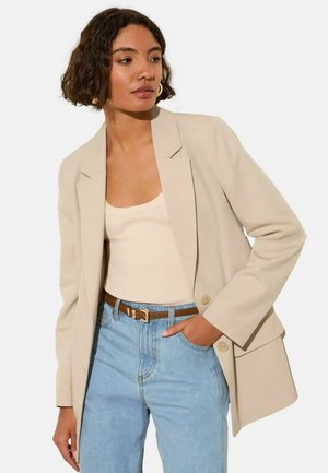 Beige blazer with a single row of buttons, notch lapels, and relaxed fit. Worn over a light tank top with blue high-waisted jeans.