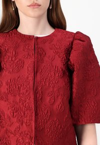 Red textured jacket featuring floral embroidery, a round neck, and short puffed sleeves. Closed with a central zipper.