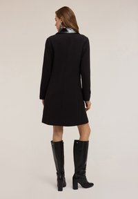 Abrigo negro con cuello de cuero negro brillante, con una silueta recta, mangas largas y largo hasta medio muslo. Combinado con botas negras hasta la rodilla.
