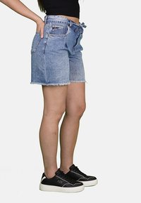 Shorts in denim blu con orlo sfrangiato, tasche frontali e vita regolabile con fiocco. Indossati con sneaker nere con suole bianche e un marchio discreto.