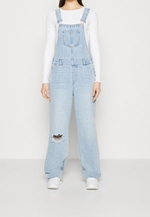 Jasnoniebieskie ogrodniczki denimowe o luźnym kroju, z regulowanymi ramiączkami, przednią kieszenią i przetarciami na kolanach. Połączone z białą bluzką z długim rękawem.