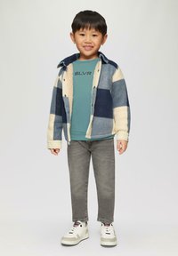 Kind met een marineblauwe en crèmekleurige geruite fleece jack over een teal sweatshirt met de tekst "SLVR" erop gedrukt. Grijze jeans en witte sneakers maken de outfit compleet.