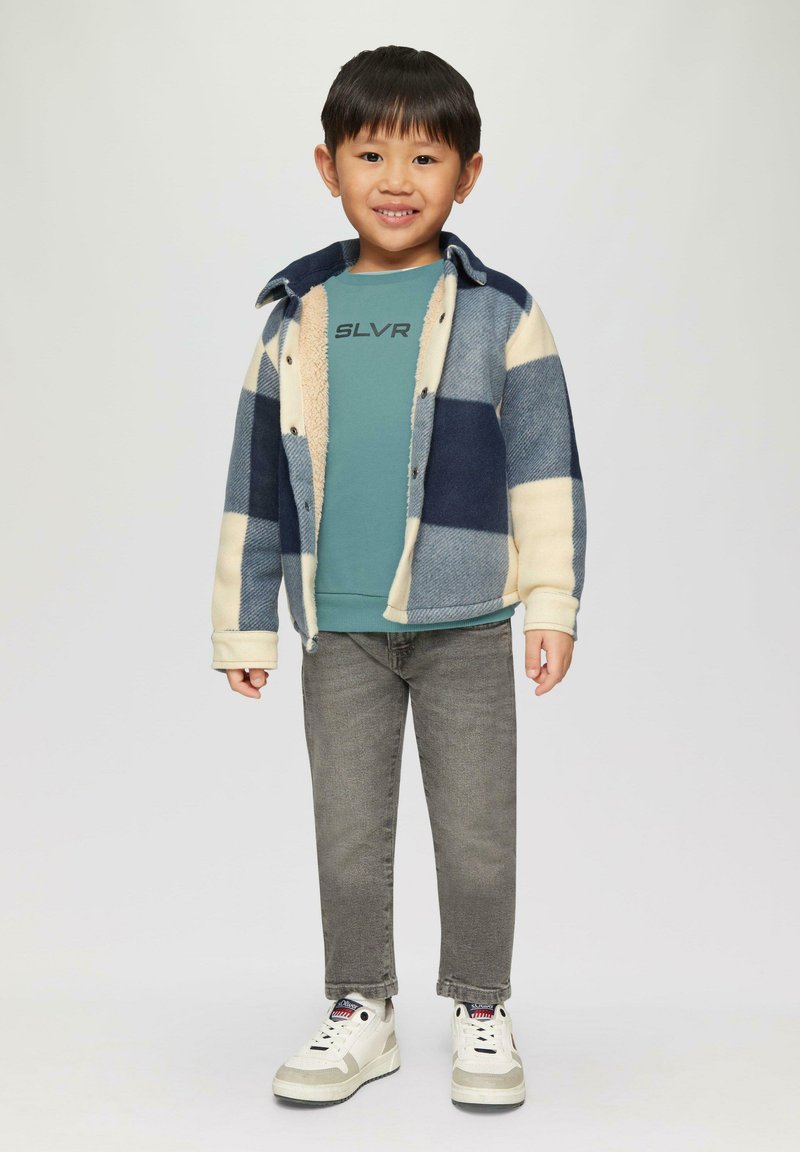 Kind met een marineblauwe en crèmekleurige geruite fleece jack over een teal sweatshirt met de tekst "SLVR" erop gedrukt. Grijze jeans en witte sneakers maken de outfit compleet.