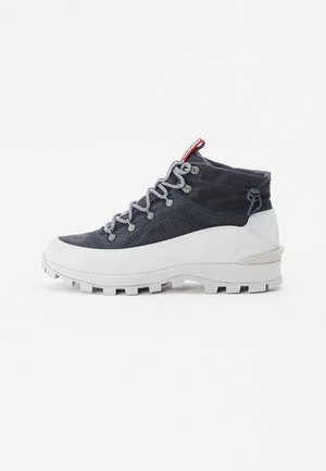 Botas de senderismo de caña alta con una parte superior texturizada en azul oscuro, suela blanca, cordones blancos en contraste y un acento rojo en el talón.