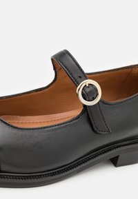 Zwarte leren schoen met een ronde neus en een kleine hak. Heeft een bandje met een gouden accent. Gladde textuur met contrasterende bruine binnenkant.