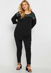 Schwarze Tunika mit floraler Stickerei am transparenten Halsausschnitt, kombiniert mit schwarzen Leggings und schwarzen Blockabsatzsandalen. Glatter Stoff.