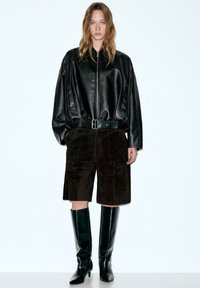 Übergroße schwarze Lederjacke mit durchgehendem Reißverschluss, kombiniert mit braunen Wildledershorts und kniehohen schwarzen Stiefeln mit Absatz. Schlichtes, modernes Design.