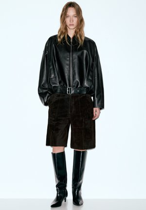 Fekete, extra méretű bőrkabát zipzáras elülső résszel, barna velúr rövidnadrággal és térdig érő fekete, sarkú csizmával párosítva. Egyszerű, modern dizájn.