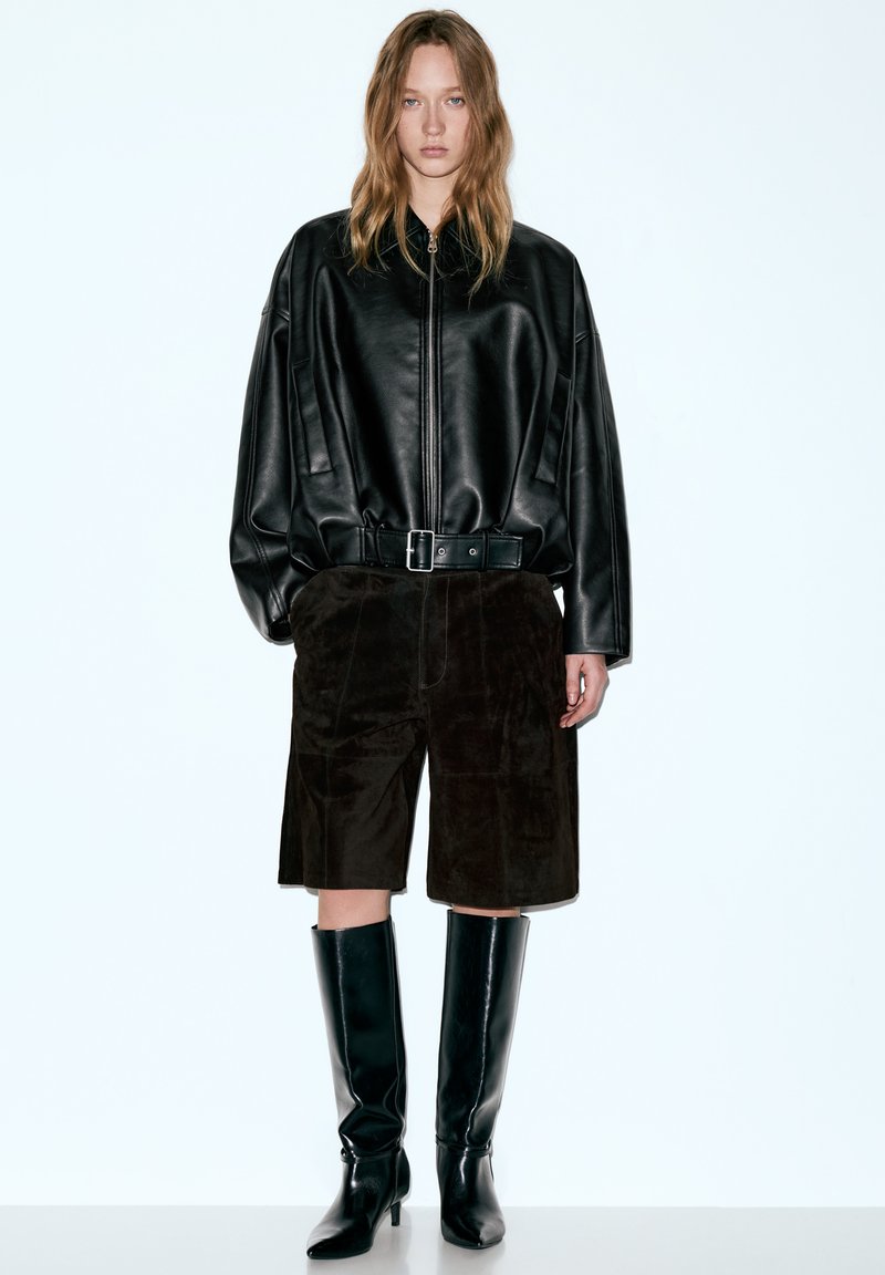 Übergroße schwarze Lederjacke mit durchgehendem Reißverschluss, kombiniert mit braunen Wildledershorts und kniehohen schwarzen Stiefeln mit Absatz. Schlichtes, modernes Design.