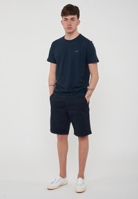 T-shirt navy a collo alto con un piccolo logo, abbinato a pantaloni corti navy. Modello che indossa sneakers bianche, in piedi con le mani in tasca.