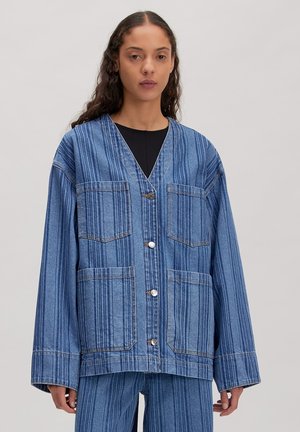 Frau trägt lockere blaue Jeansjacke und passende Hosen mit vertikalen Streifen vor einfarbigem Hintergrund.