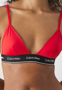 Rød trekantet bralette laget av glatt stoff, med tynne stropper og et svart elastisk bånd med hvit "Calvin Klein" tekst.