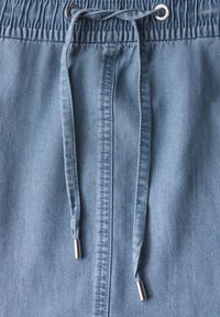 Ljusblå denimtyg har en elastisk midja med dragsko och metallaglets, samt vertikala sömnadsdetaljer överallt.