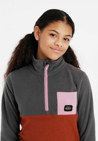 Pull en polaire gris avec un col zippé, poche poitrine rose et section inférieure orange. Présente un patch logo noir sur la poche.