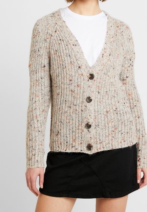 Beige neuleliivi, jossa on v-pääntie, pitkät hihat ja viisi tummaa nappia. Kankaassa on oransseja ja ruskeita sävyjä, jotka tuovat tekstuuriin vivahteita.