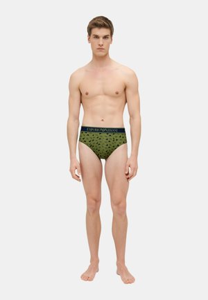 Slips verdi con motivo a fantasia e una cintura blu navy con il marchio, caratterizzati da un design geometrico. Il materiale sembra morbido e elasticizzato. Il modello è in piedi a piedi nudi.