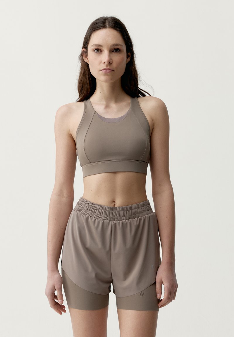Young woman with long dark hair wearing light brown sports bra and matching layered shorts, standing against plain background.