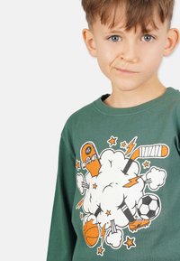 Grön långärmad skjorta med en grafisk design av en skateboardåkare, sportutrustning, blixtar och stjärnor i lila orange och vitt.