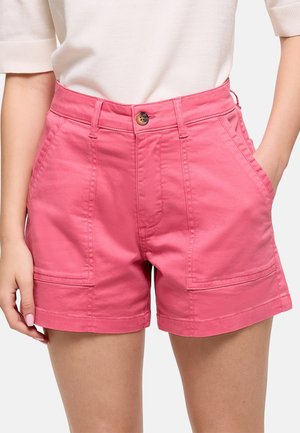 Mujer vistiendo shorts de algodón rosa de cintura alta con bolsillos delanteros y cierre de botones, combinados con una camiseta blanca de manga corta.