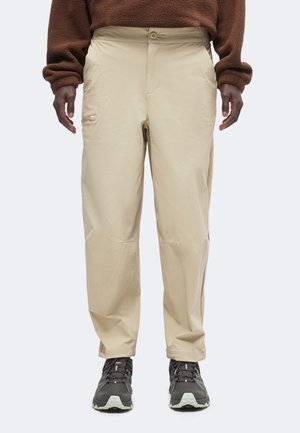 Persona con pantalones beige, zapatillas oscuras y una camiseta marrón de manga larga, de pie frente a un fondo liso.