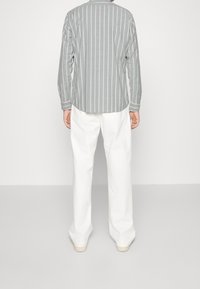 Szara i biała koszula w paski z długim rękawem i zaokrąglonym dołem, w zestawieniu z luźnymi białymi spodniami, na neutralnym tle.